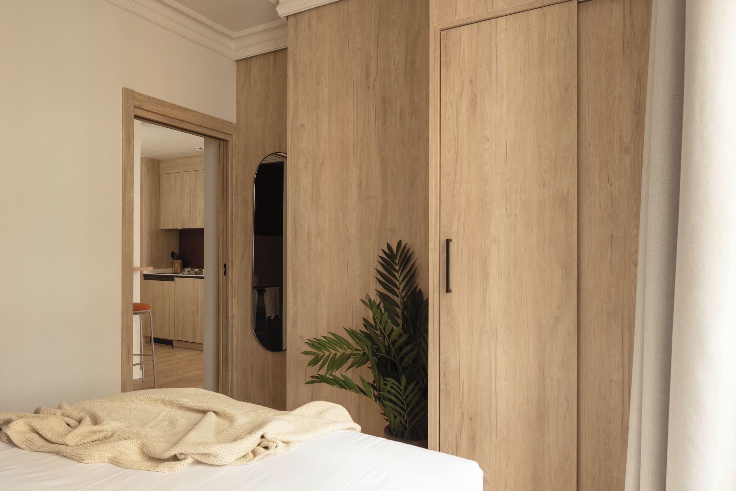 A bedroom with a bed, a wooden wardrobe, a mirror, and a potted plant.