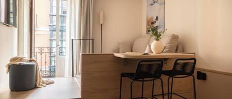 A modern kitchen with a wooden countertop, two black bar stools, a small round table with a beige cushion, and a vase with flowers.