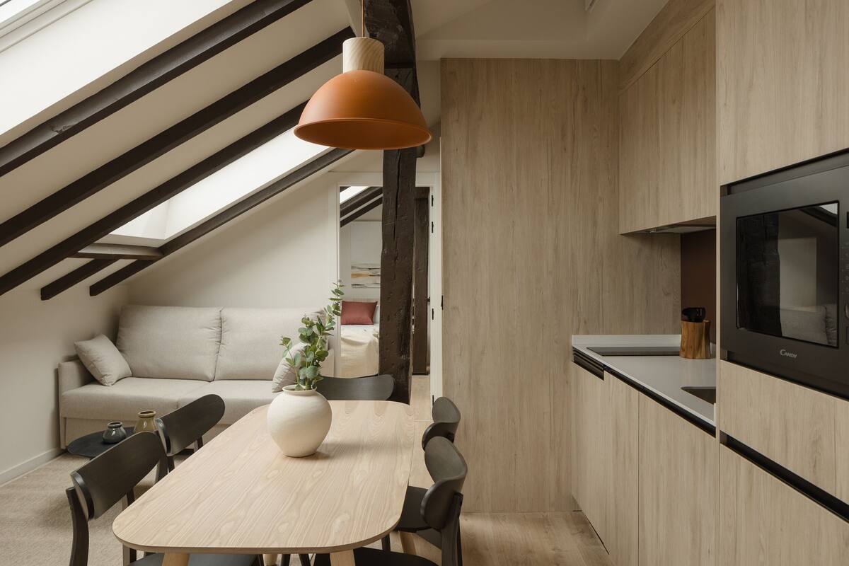 A modern kitchen with a dining area, featuring a wooden table, chairs, a large vase, and a TV mounted on the wall.