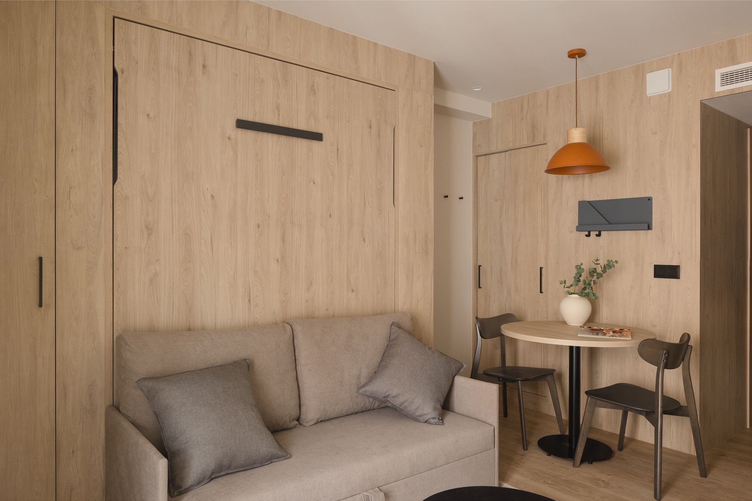 Een moderne kamer met een houten garderobe, een bank met kussens, een ronde tafel met stoelen en een hangende hanglamp.