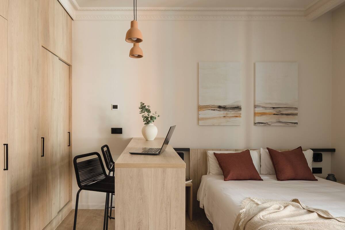 A bedroom with a bed, a desk with a laptop, a chair, and a plant.
