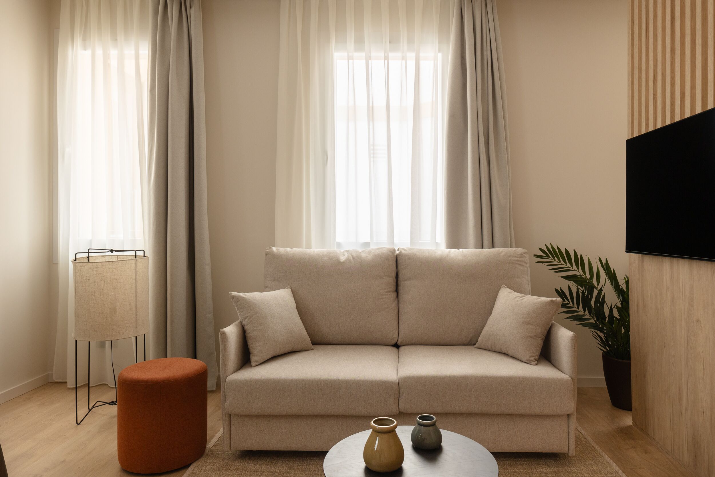 Una sala de estar moderna con un sofá color beige, un otoman redondo y una pequeña mesa con dos jarrones.