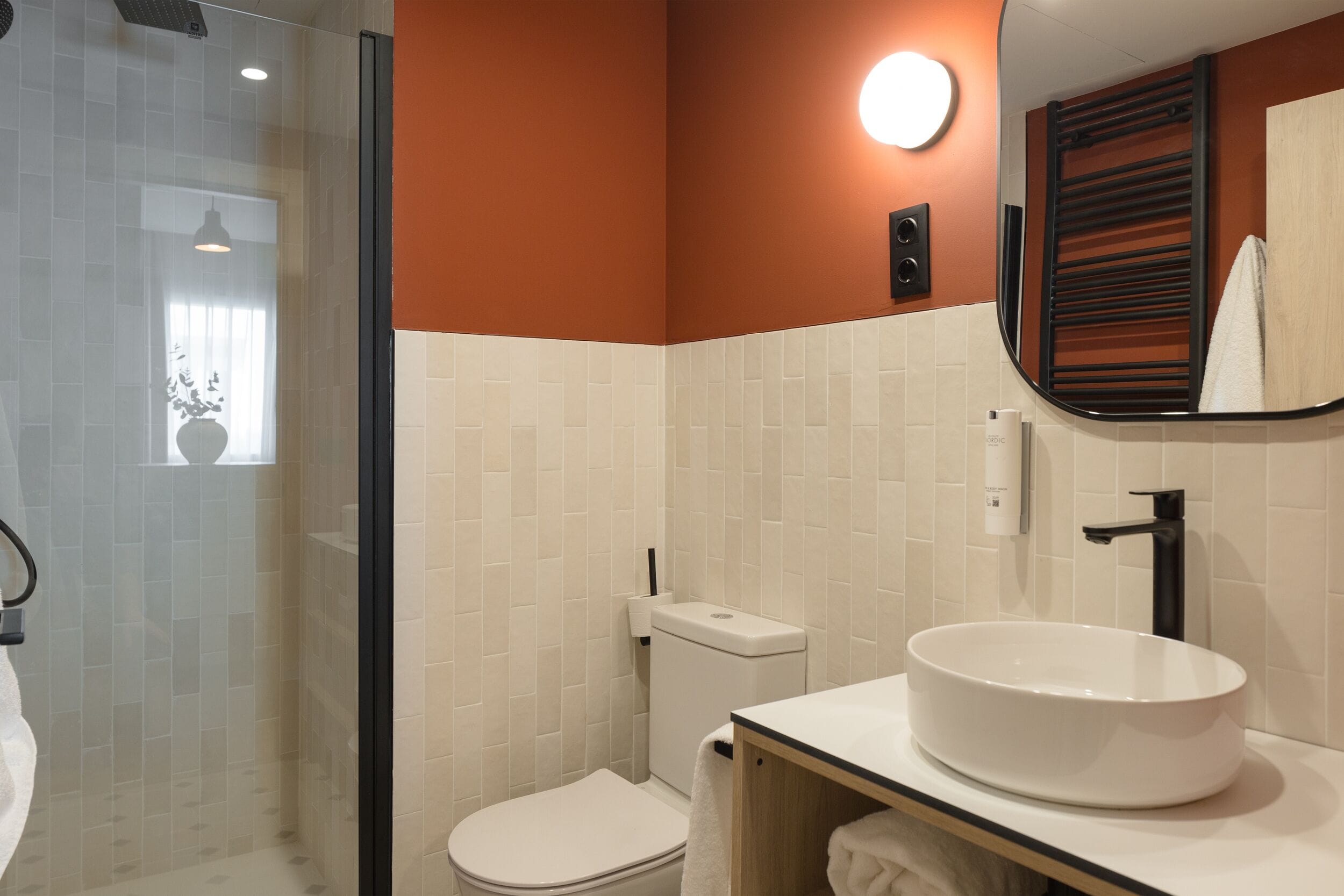 Een badkamer met een glazen douchecabine, een wit toilet, een rond wit wastafeltje en een spiegel met houten lijst.