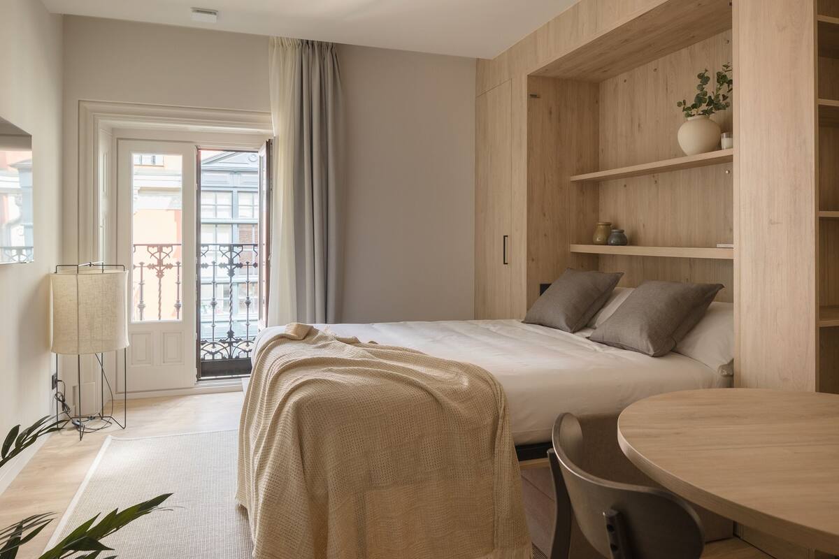 A modern bedroom with a large bed, a wooden wardrobe, a dining table, and a balcony with a view.