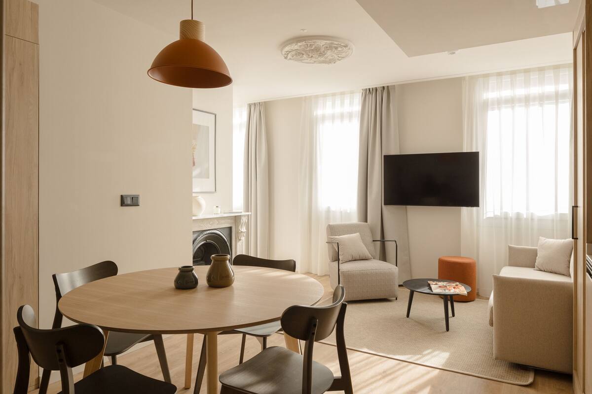 A modern living room with a round wooden table, several chairs, a sofa, a TV, and a fireplace.