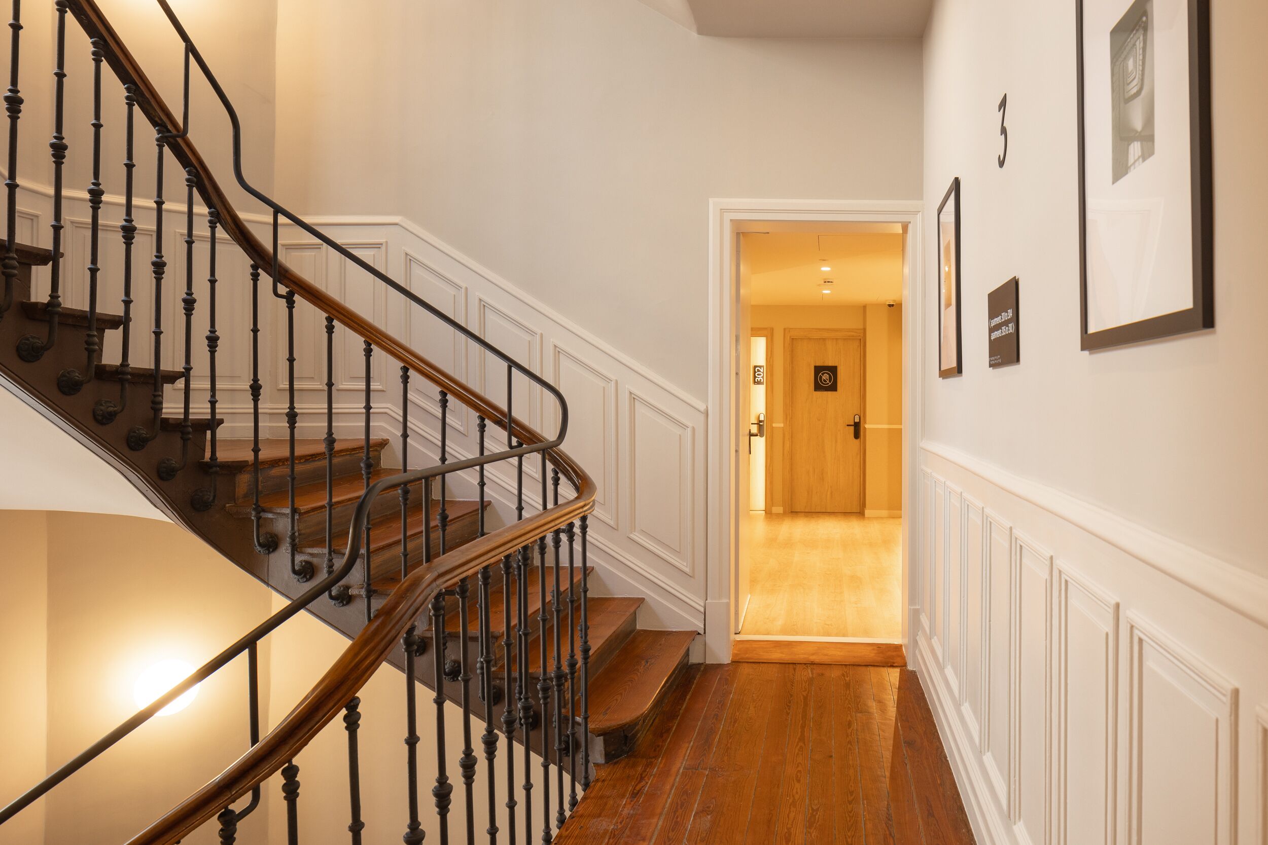 A staircase with a wooden handrail and black balusters, leading to a room with a wooden door and framed pictures on the wall.