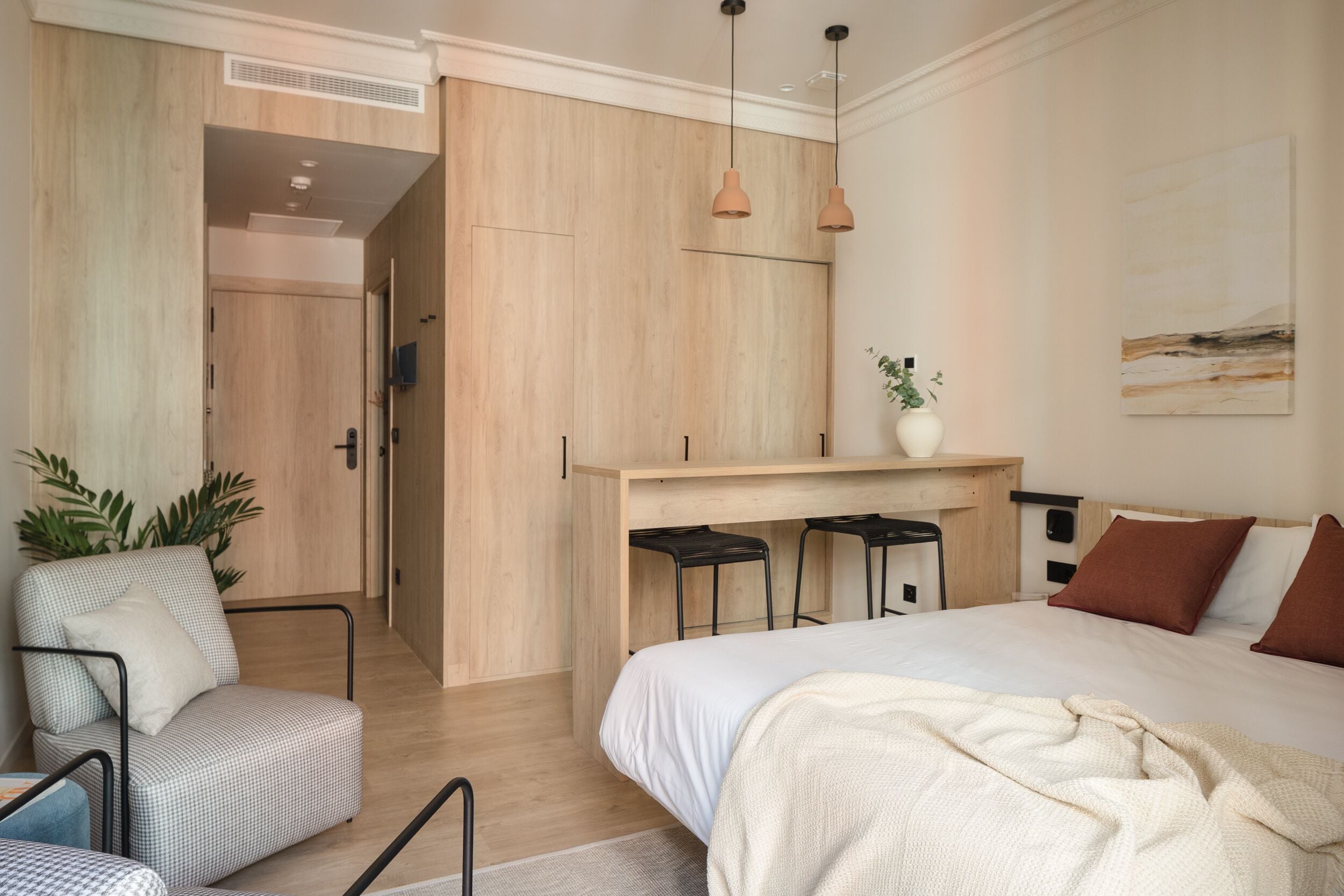 Een moderne slaapkamer met een groot bed, een bureau met twee krukken, een stoel en een plant.
