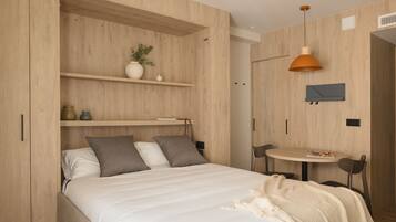 Un dormitorio moderno con una cama, una mesita pequeña y una silla. Encima de la cama hay una estantería con objetos decorativos.