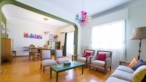 Un salon spacieux doté d’un parquet en bois, d’une table basse verte et d’un canapé agrémenté de coussins colorés. On aperçoit au fond deux fauteuils et une table à manger avec des chaises.