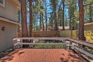 Une terrasse en bois avec une balustrade blanche, entourée de grands pins, mène à un bâtiment doté d’un toit à pignon.