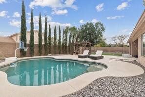 A swimming pool with a curved edge, surrounded by a pebble border. There are lounge chairs and a pergola with a roof. Tall trees are visible in the background. - Hues of the Horizon House with Pool & BBQ (Scottsdale)