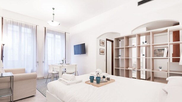 A modern hotel room with a large bed, a desk, a chair, and a bookshelf.