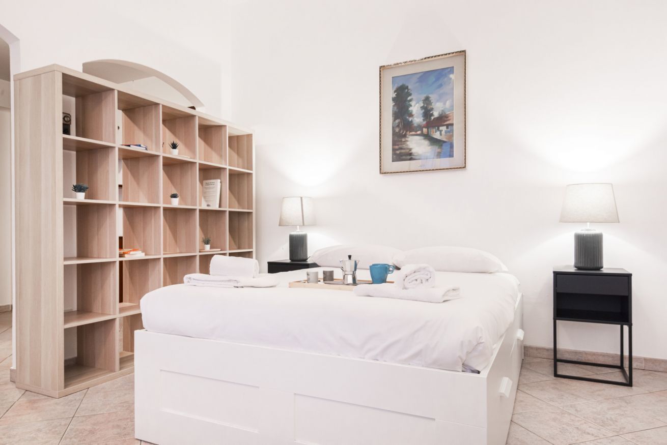 A bedroom with a white bed, a wooden shelving unit, two bedside tables with lamps, and a framed painting on the wall.
