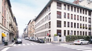 A street view with a row of parked cars, a pedestrian crossing, and buildings with multiple stories and windows.