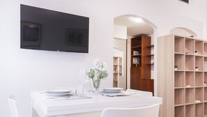 A dining area with a white table set for two, a flat-screen TV mounted on the wall, and wooden shelving units.