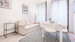 A modern living room with a sofa, a glass coffee table, a dining table with chairs, and a patterned rug.
