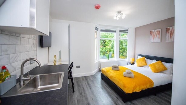 Ein modernes Hotelzimmer mit Küchenzeile, einem großen Fenster und Blick ins Grüne sowie einem Bett mit gelber Bettwäsche.