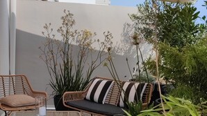An outdoor seating area with wicker furniture and a glass table, surrounded by greenery and a white wall. - La Suite Appartement Avec Terrasse Proche Plages Tout a Pied (Concarneau)