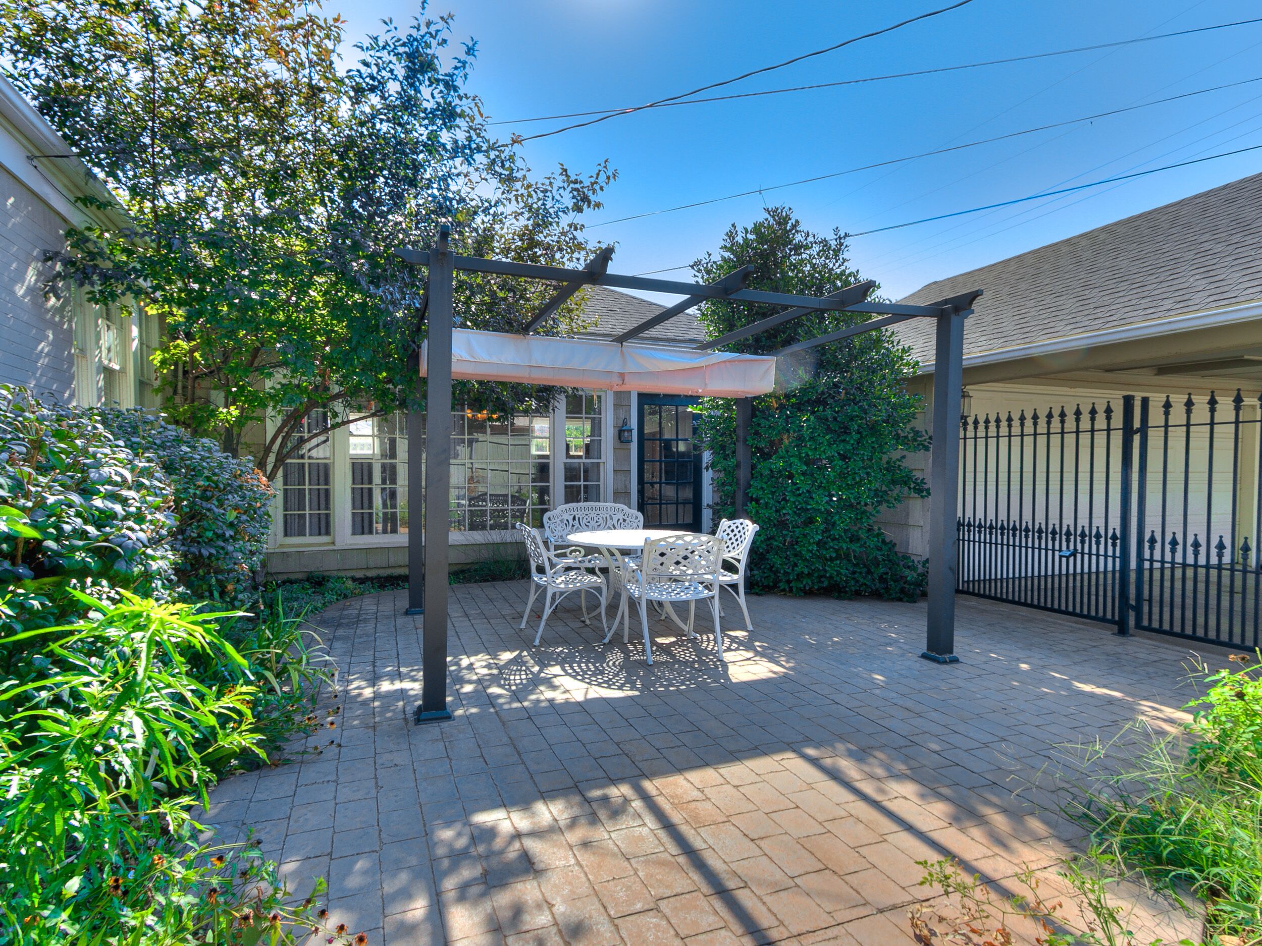 A patio with a table and chairs under a pergola, surrounded by greenery and a brick pathway.
