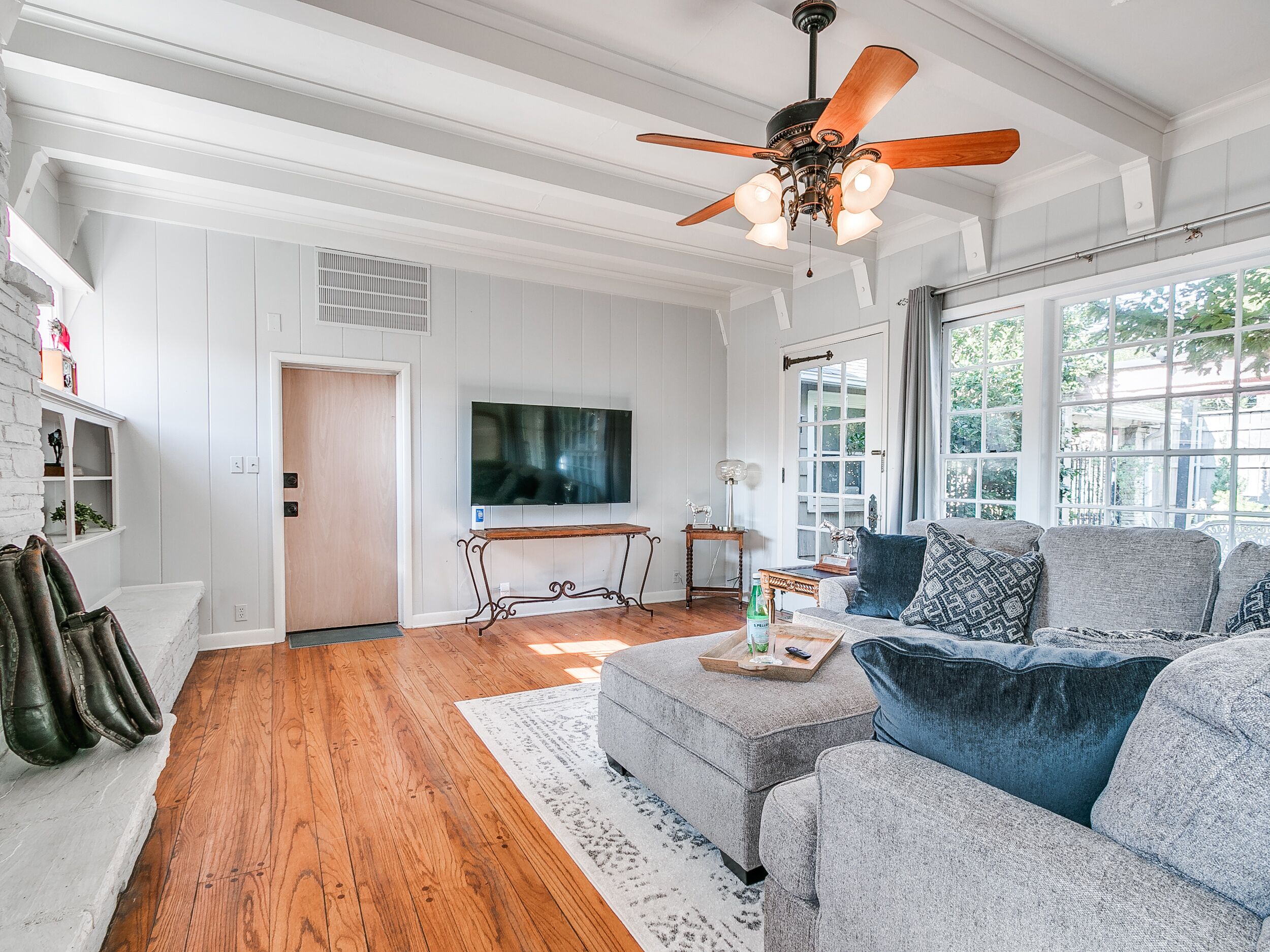 A spacious living room with a wooden floor, a ceiling fan, a large flat-screen TV, and a seating area with grey sofas and a coffee table.