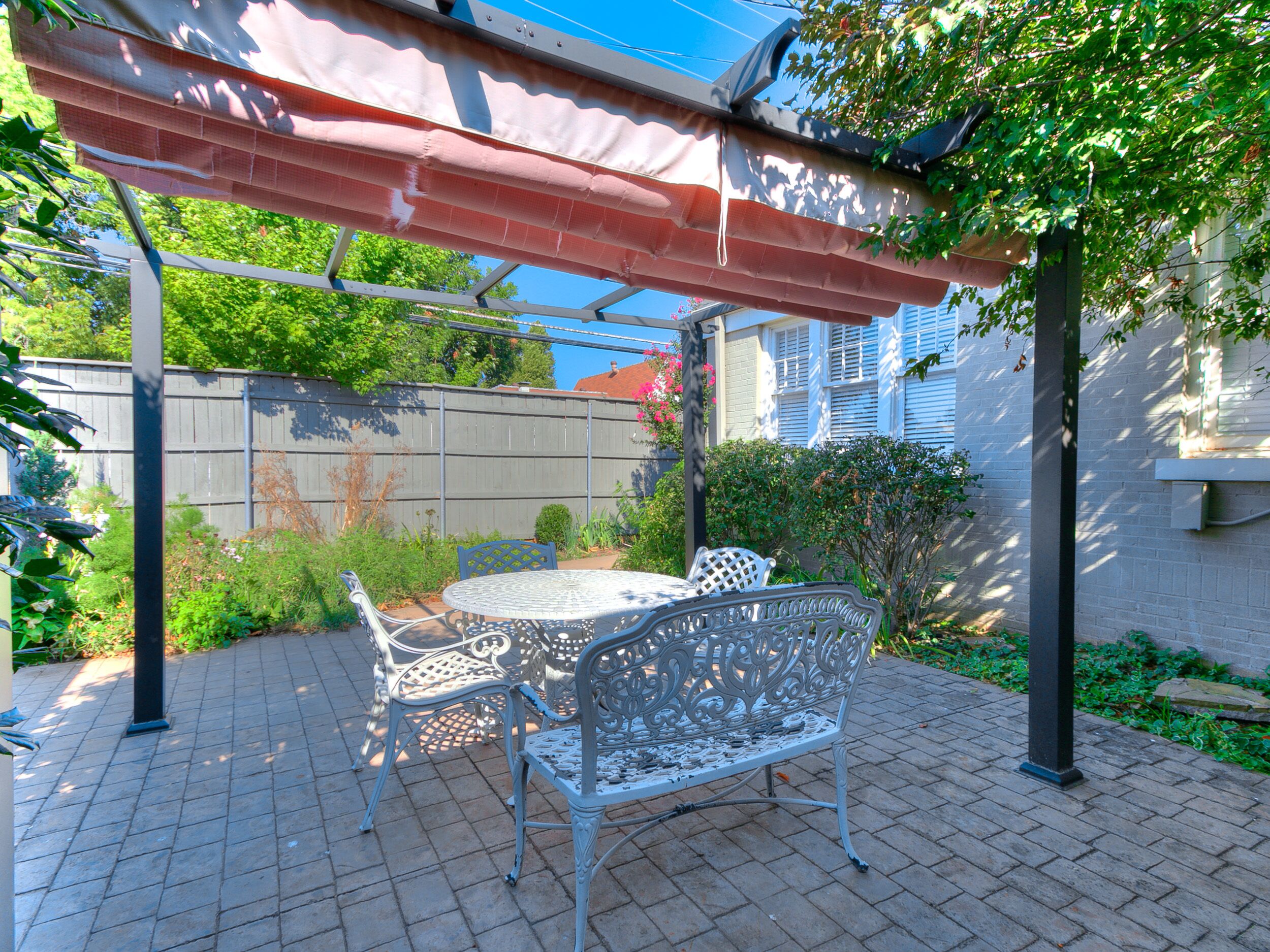 Une terrasse avec une table et des chaises en métal, une pergola surmontée d’une toile rouge et, au fond, un mur de briques.