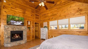 Une chambre située dans un chalet en rondins, avec une cheminée en pierre, une télévision, un lit et une fenêtre donnant sur le paysage extérieur.