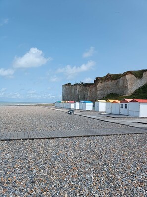 A pebbled beach with a wooden pier, a cliff, and colorful beach huts.