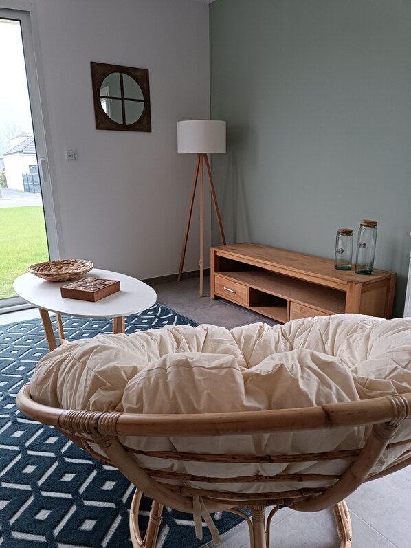 Ein Zimmer mit einem Korbstuhl, einem weißen Tisch, einer Lampe, einem Holzschrank und Blick ins Freie.