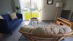 Olohuoneessa on sininen sohva, rottinkituoli, pieni tarjotinpöytä, televisio puisen jalustan päällä ja suuret ikkunat, joista on näkymä ulos.