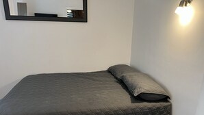 En enkeltseng med en quiltet madras og en pude, en dør med håndtag og en vægmonteret lampe.