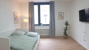 Een slaapkamer met een bed, een raam met gordijnen, een radiator, een potplant en een aan de muur bevestigde televisie.
