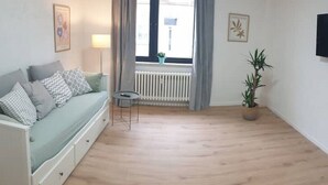 Een kamer met een lichtbeige bank, een klein rond tafeltje, een potplant en een raam met gordijnen.