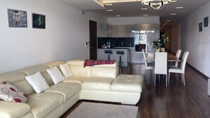 Una sala moderna con un sofá seccional color beige, un comedor con una mesa y sillas blancas, y una cocina al fondo.