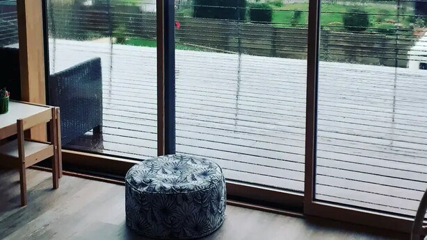 Ein Zimmer mit einer großen Schiebeglasdoor, die auf einen Außenbalkon führt. Der Balkon hat ein Metallgeländer und einen Holzboden. Auf dem Boden steht ein runder Hocker mit Blumenmuster.