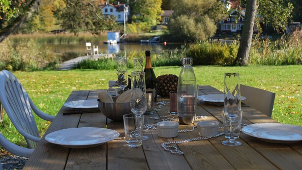 Een houten tafel gedekt voor een maaltijd, compleet met borden, glazen en een fles, met uitzicht op een meer met huizen op de achtergrond.