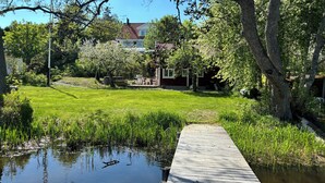 Een rustig pad langs de rivier dat leidt naar een huis met een rood dak, omgeven door weelderige begroeiing en bomen.
