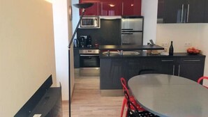 A modern kitchen with dark cabinetry, a stainless steel refrigerator, a microwave, and a coffee maker. There is a dining area with a round table and red chairs.