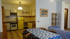 Eine kleine Küche mit gelbem Kühlschrank, Herd und Holzschränken. Ein Esstisch mit Stühlen und einer blauen Tischdecke steht bereit. Ein Bild hängt an der Wand über dem Essbereich.