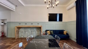 A modern living room with a dining table, clear chairs, a blue sofa, a fireplace, and a large TV. - Fuori dal Porto2-Quadrupla VistaMare byBeYourHome (Civitavecchia)