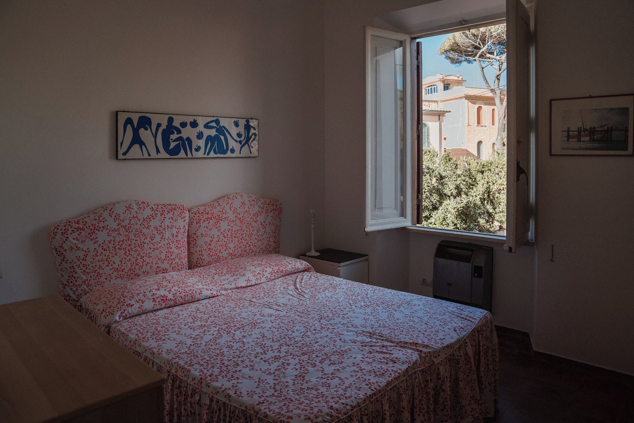 Une chambre à coucher comprenant un lit, une table de chevet, une fenêtre donnant sur des immeubles et un mur orné d’une œuvre d’art.
