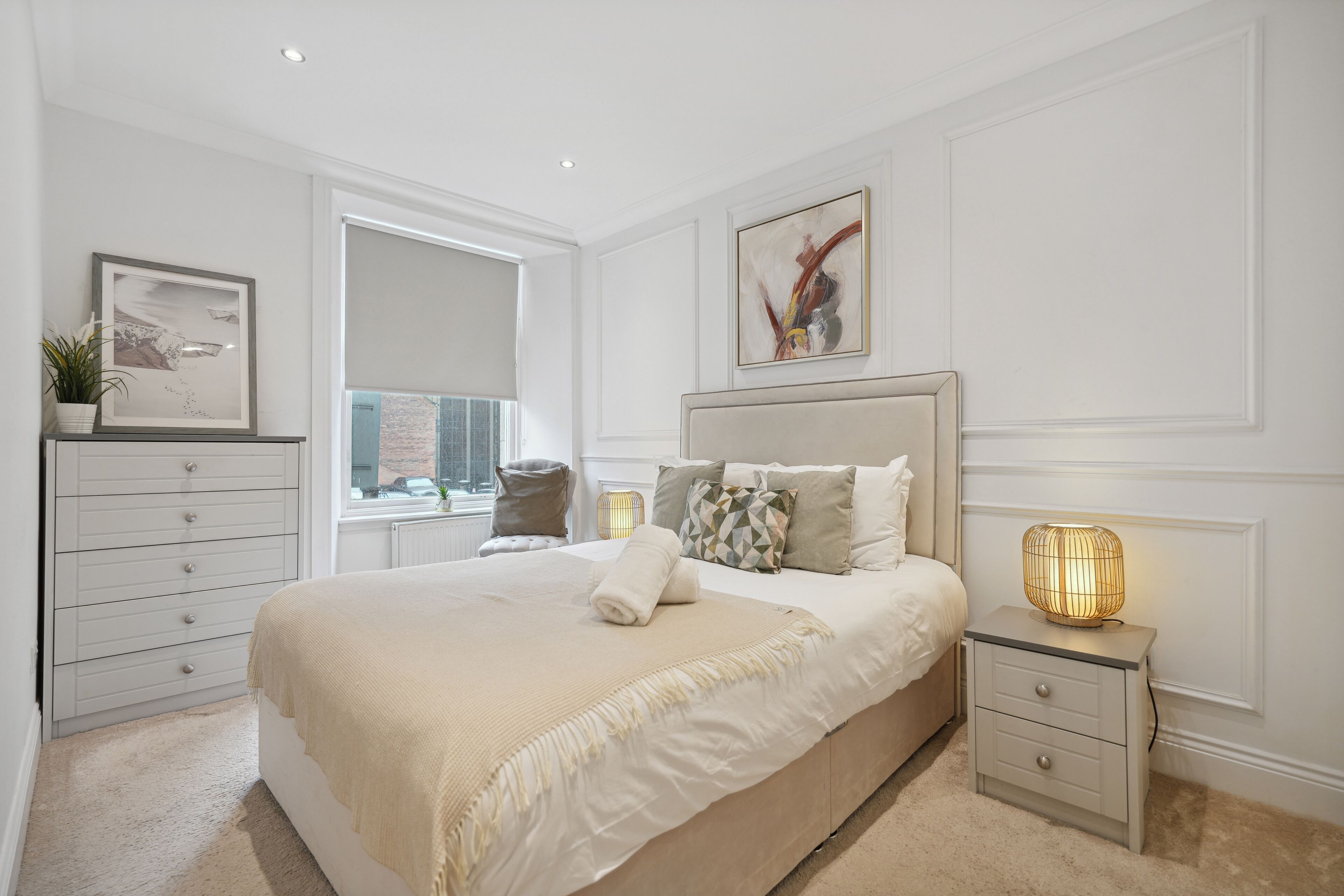 A bedroom with a large bed, a bedside table with a lamp, a dresser, and a window with a view of the city.