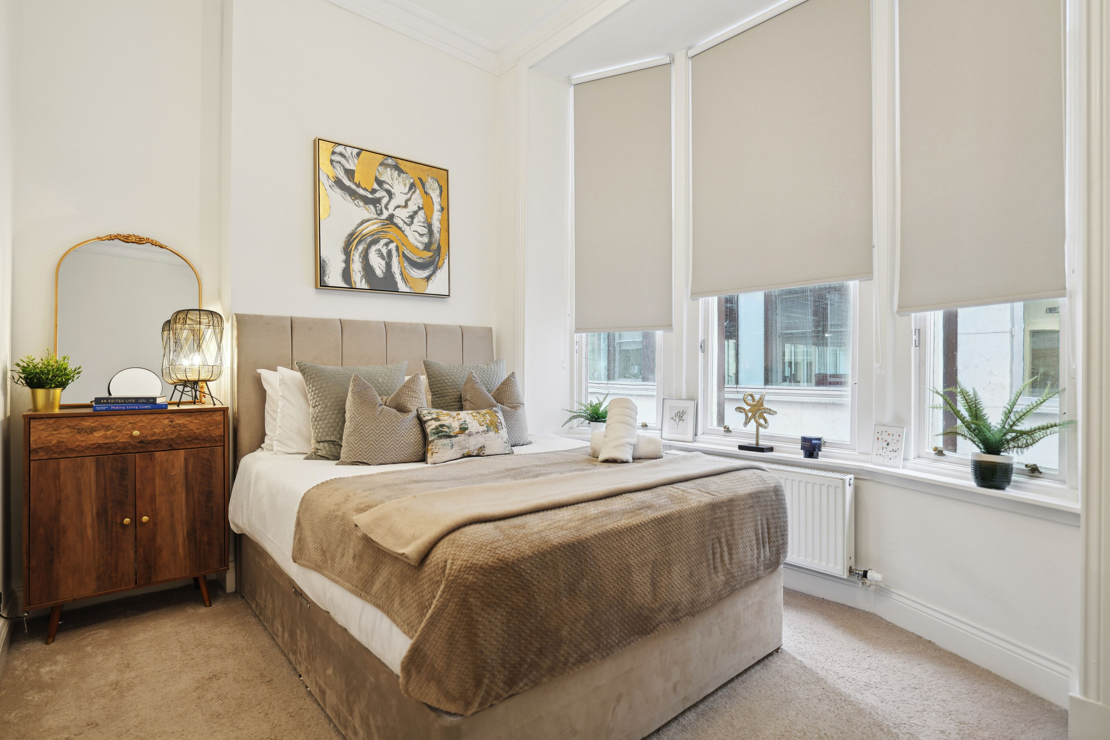 A bedroom with a large bed, a nightstand, a mirror, a painting, and a window with blinds.