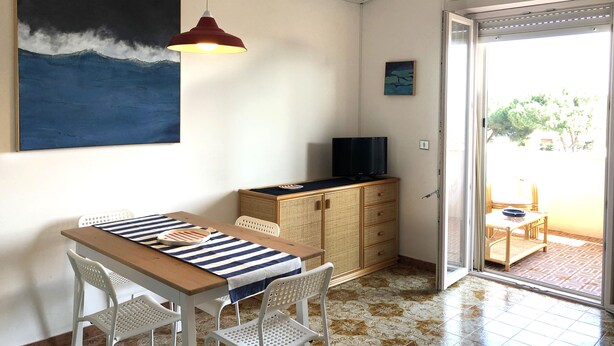 A room with a dining table, chairs, a television on a cabinet, an air conditioner, and a painting of a wave. - Casa Raffaello - by Be Your Home (Santa Marinella)
