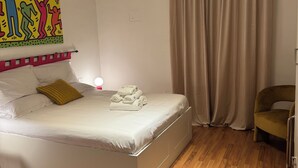 Een opgemaakt bed met witte lakens en een geel kussen, een houten vloer, een beige gordijn en een kleurrijk kunstwerk aan de muur.