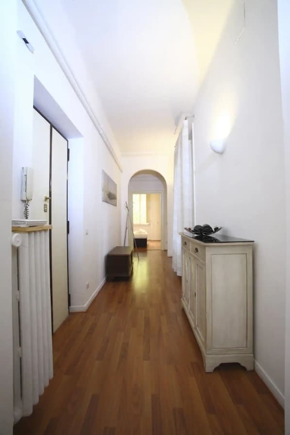 A narrow hallway with wooden flooring, white walls, a radiator, a cabinet, and a door with a telephone.