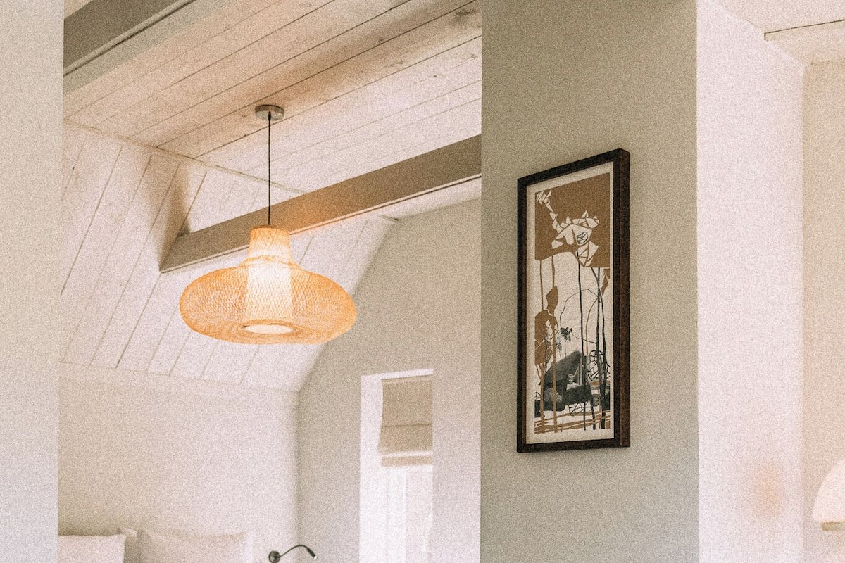Ein Schlafzimmer mit einem Bett, einer Hängelampe, einem gerahmten Bild an der Wand und einem kleinen runden Hocker.