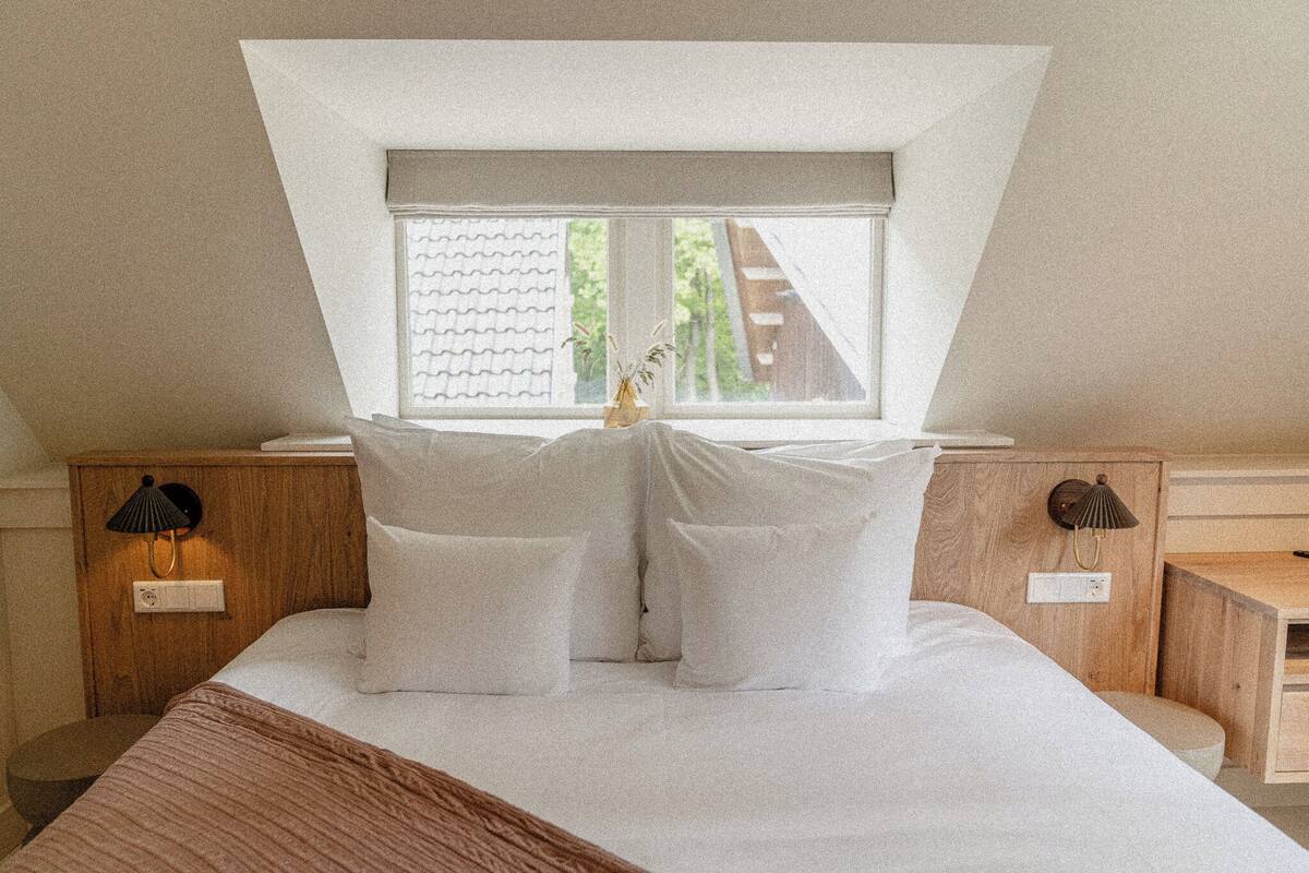 Ein Schlafzimmer mit schrägen Wänden, einem Bett mit weißer Bettwäsche, zwei Nachttischen mit Lampen und einem Fenster mit Blick ins Grüne.