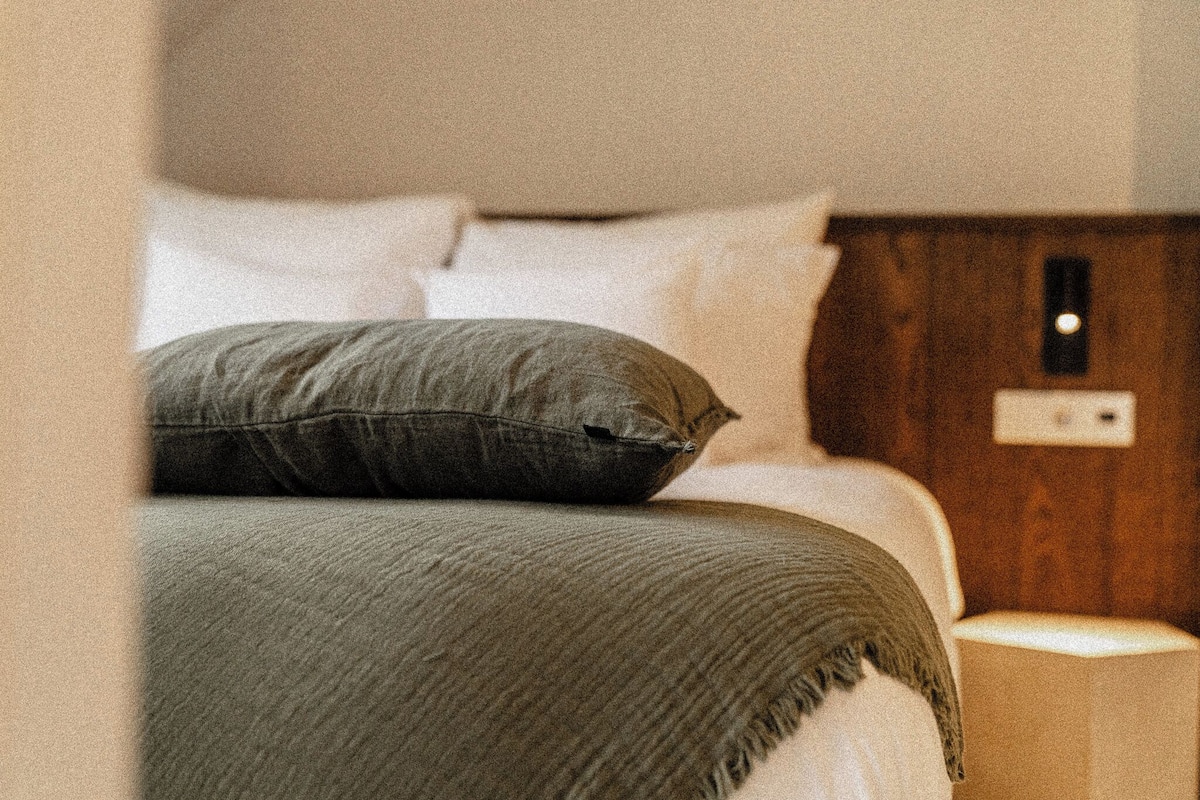 Ein ordentlich bezogenes Bett mit grüner Decke und Kissen, ein Holz-Kopfbrett sowie ein Nachttischchen mit Lampe.