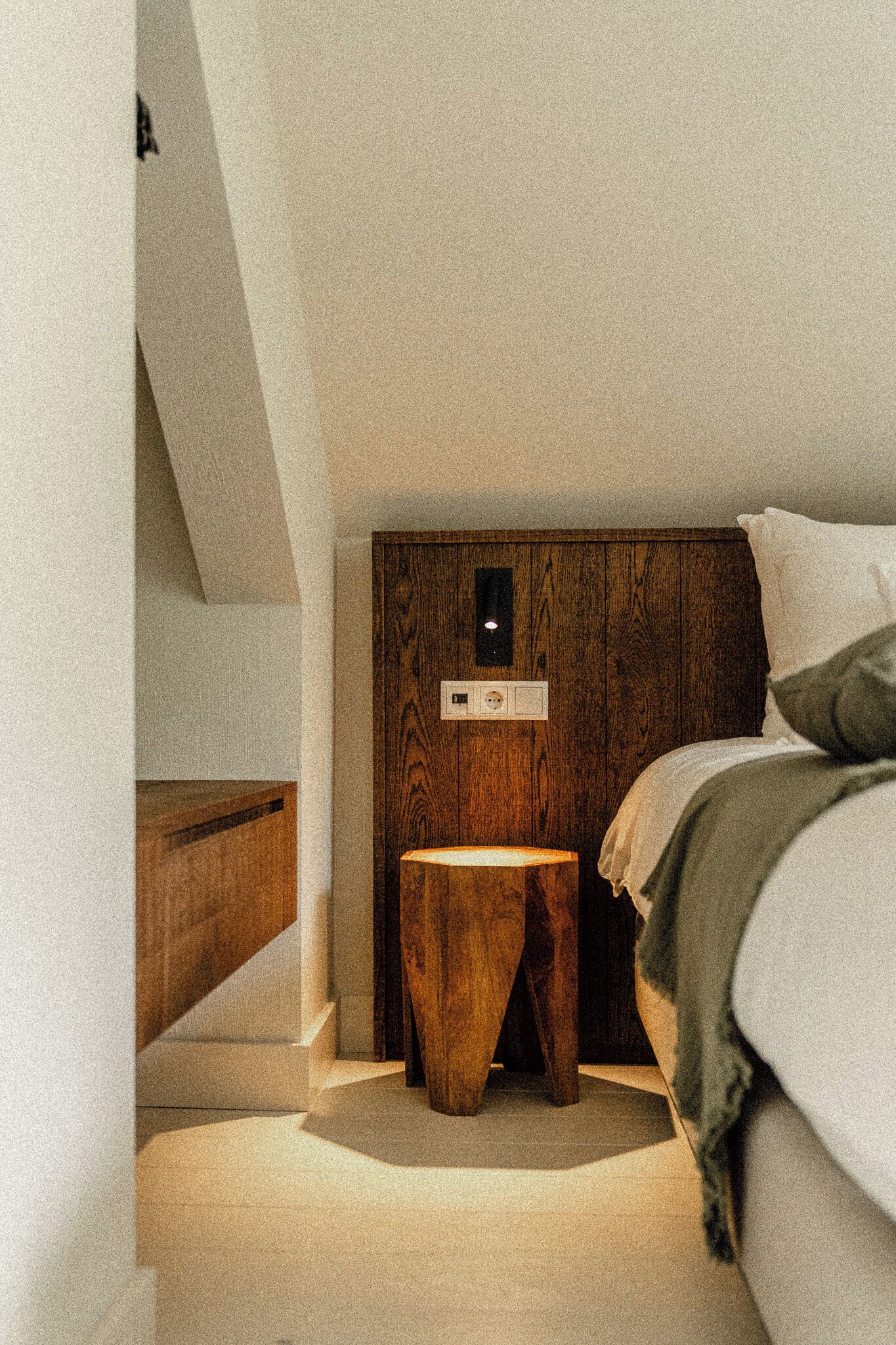Un dormitorio con techo inclinado, cabecera de madera, mesita de noche y cama con manta verde.