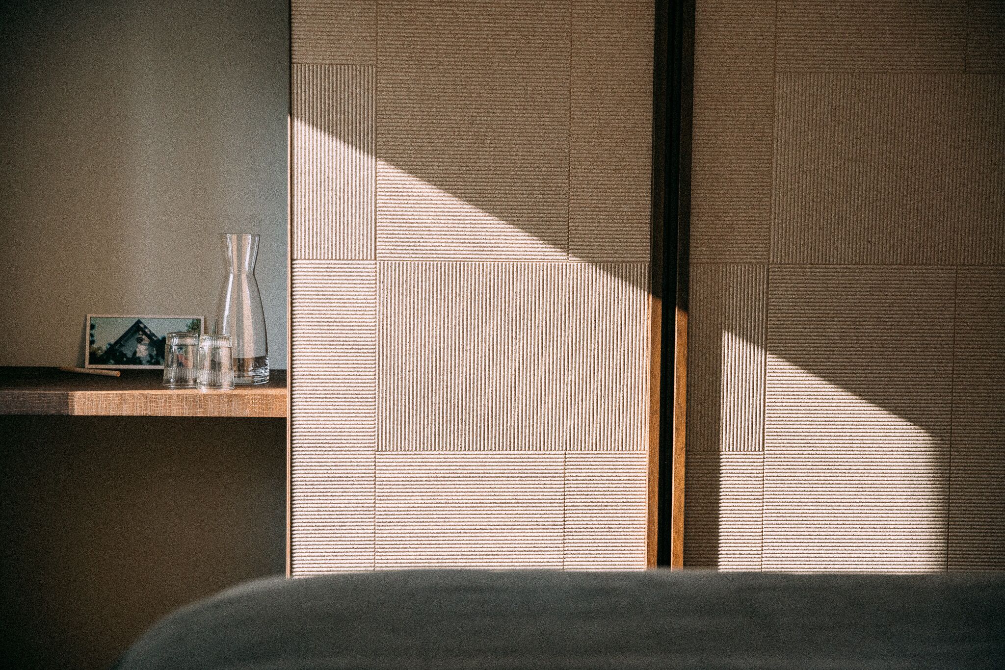Une pièce comprenant un lit, une étagère avec un vase et un cadre photo, et un mur texturé orné d’un motif de lignes verticales.